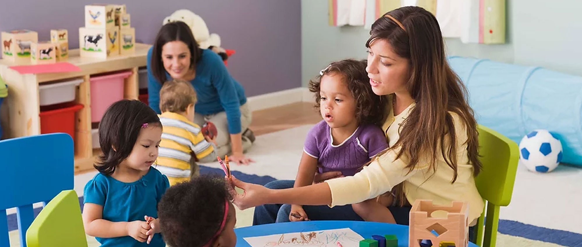 Adult engages with several young children during a classroom activity at a low table. One child sits on the adult’s lap while the adult holds up a crayon or writing tool for the children to observe. Additional children and an adult are visible in the background, along with classroom furniture and learning materials, highlighting a collaborative early childhood learning environment.