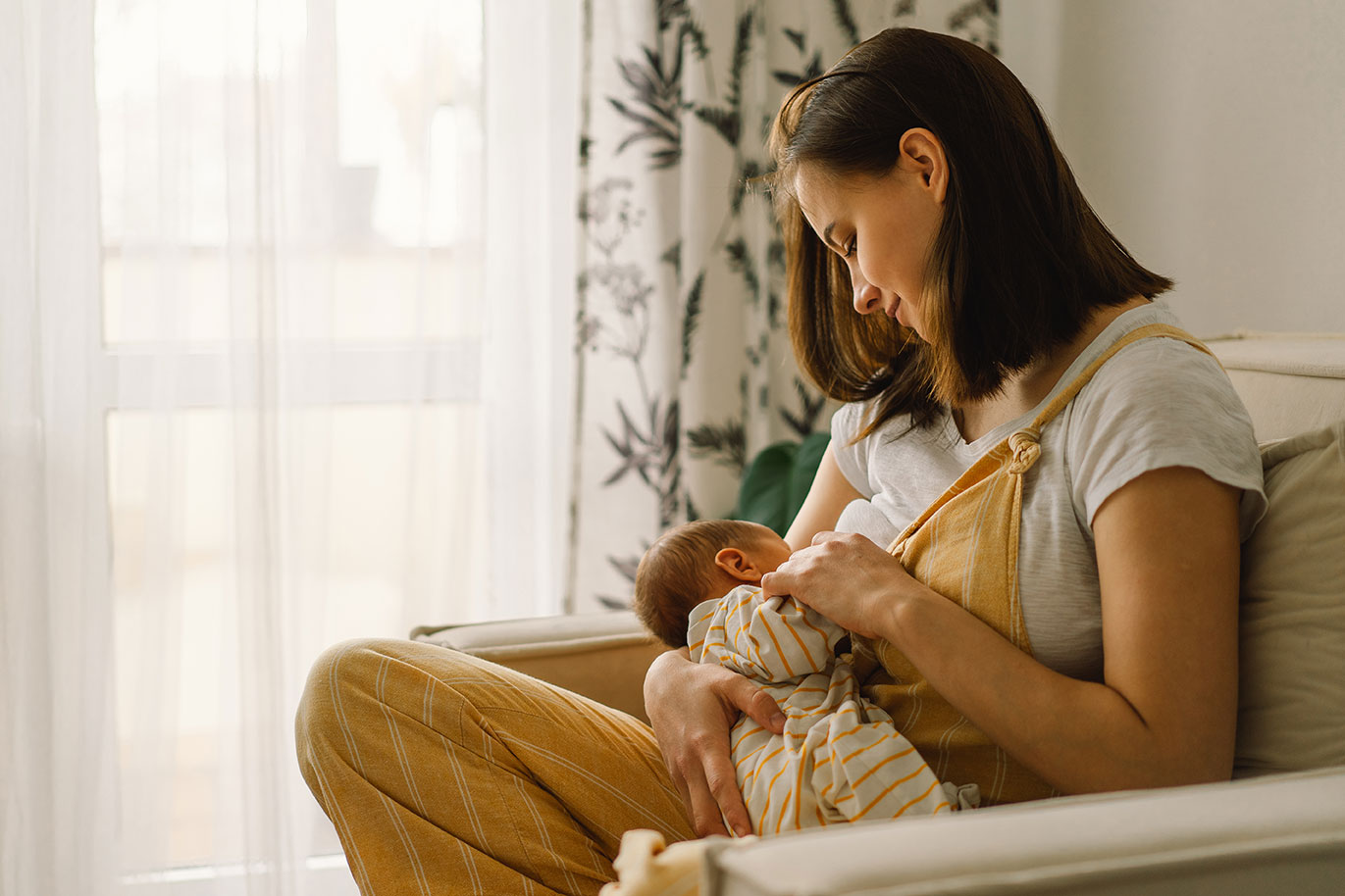 Adult seated in an armchair holding and feeding an infant in a softly lit home setting. Natural light streams through a nearby window, while patterned curtains and neutral furnishings create a calm environment. The image highlights a nurturing caregiving interaction and infant feeding.