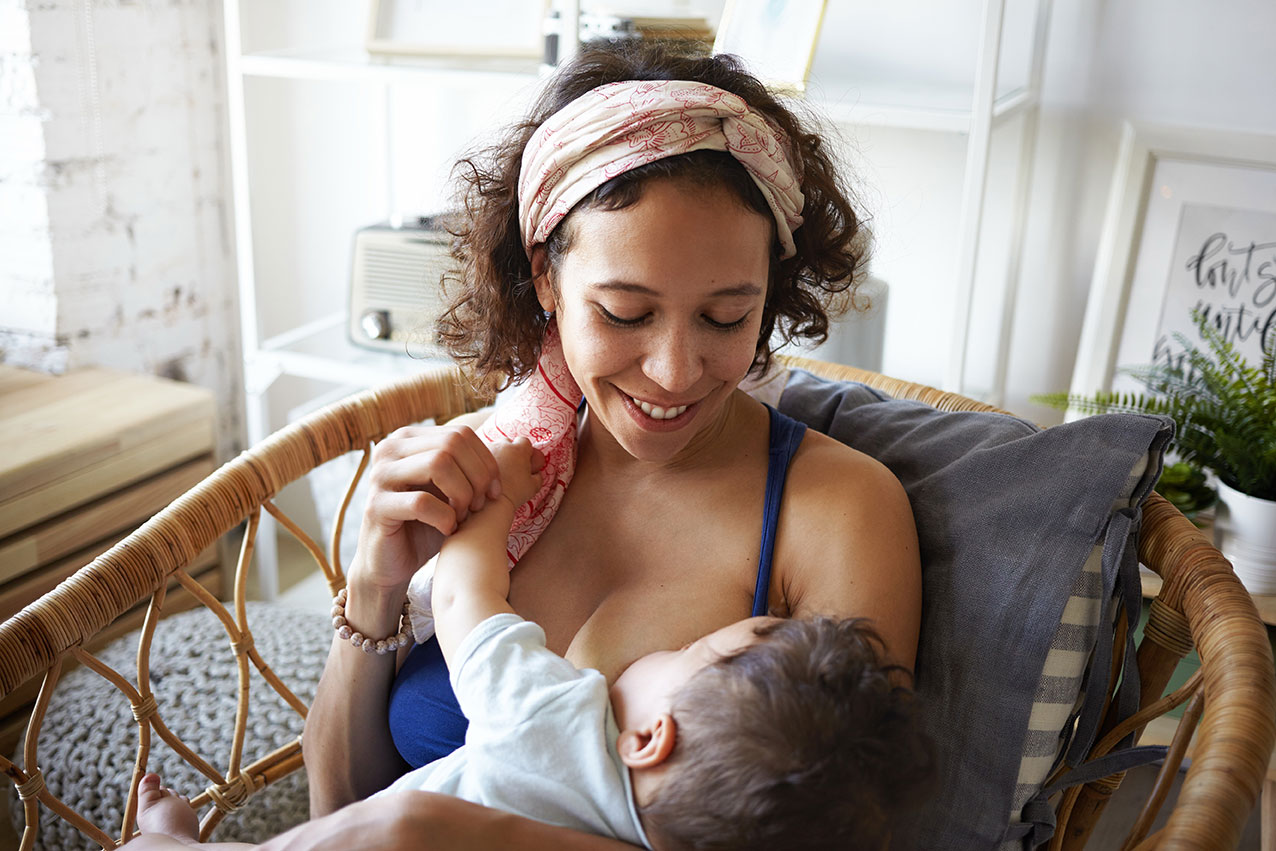 Adult seated in a wicker chair holding and feeding an infant in a home setting. The adult wears a blue top and patterned headband, while the infant rests in the adult’s arms. Shelving, framed artwork, and household furnishings are visible in the background, highlighting a nurturing caregiving interaction.