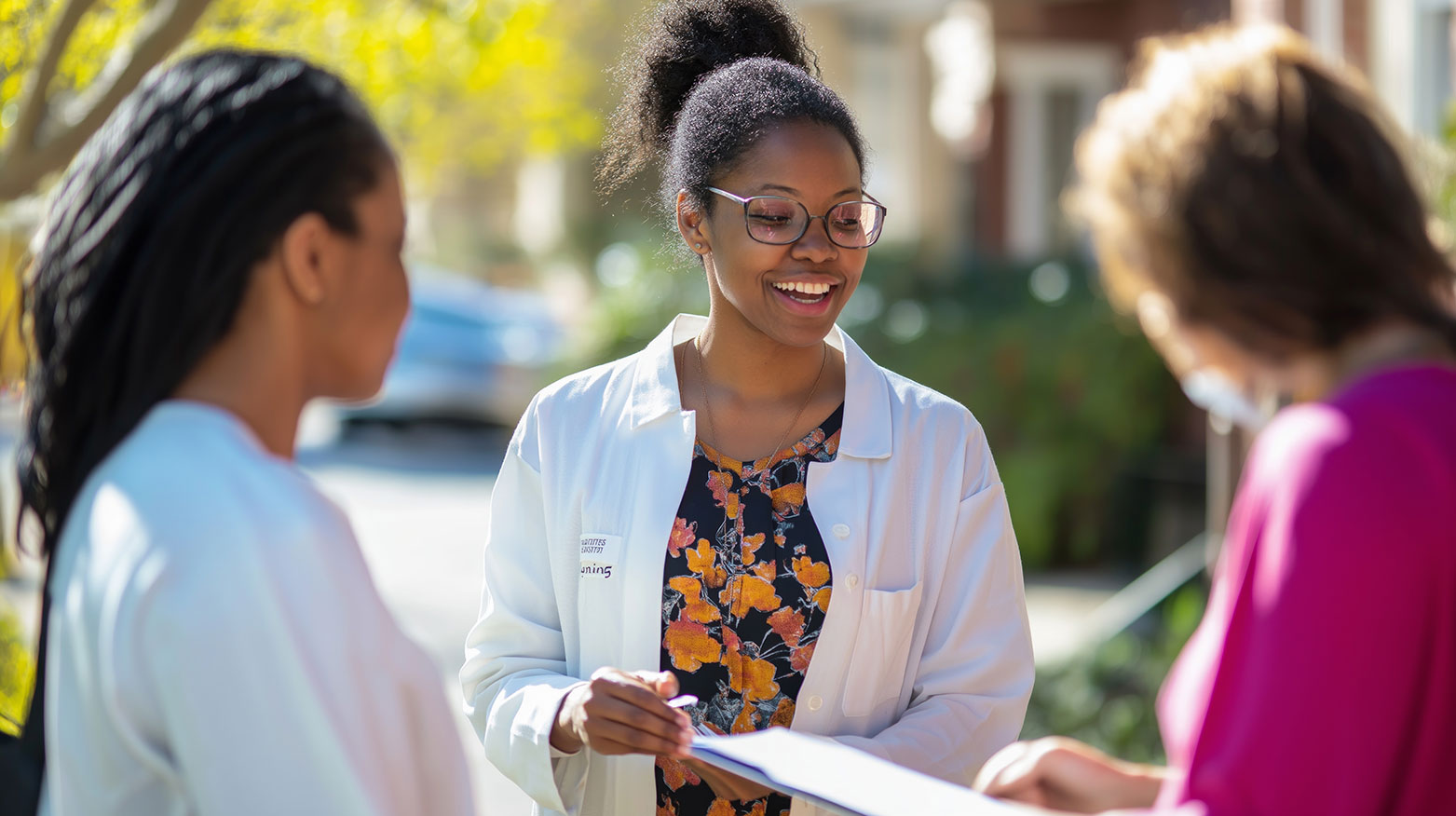 Healthcare professional wearing a white coat speaks with two individuals outdoors in a community setting. The healthcare professional holds a clipboard or notepad while standing between the two individuals, highlighting communication, guidance, and family support services.