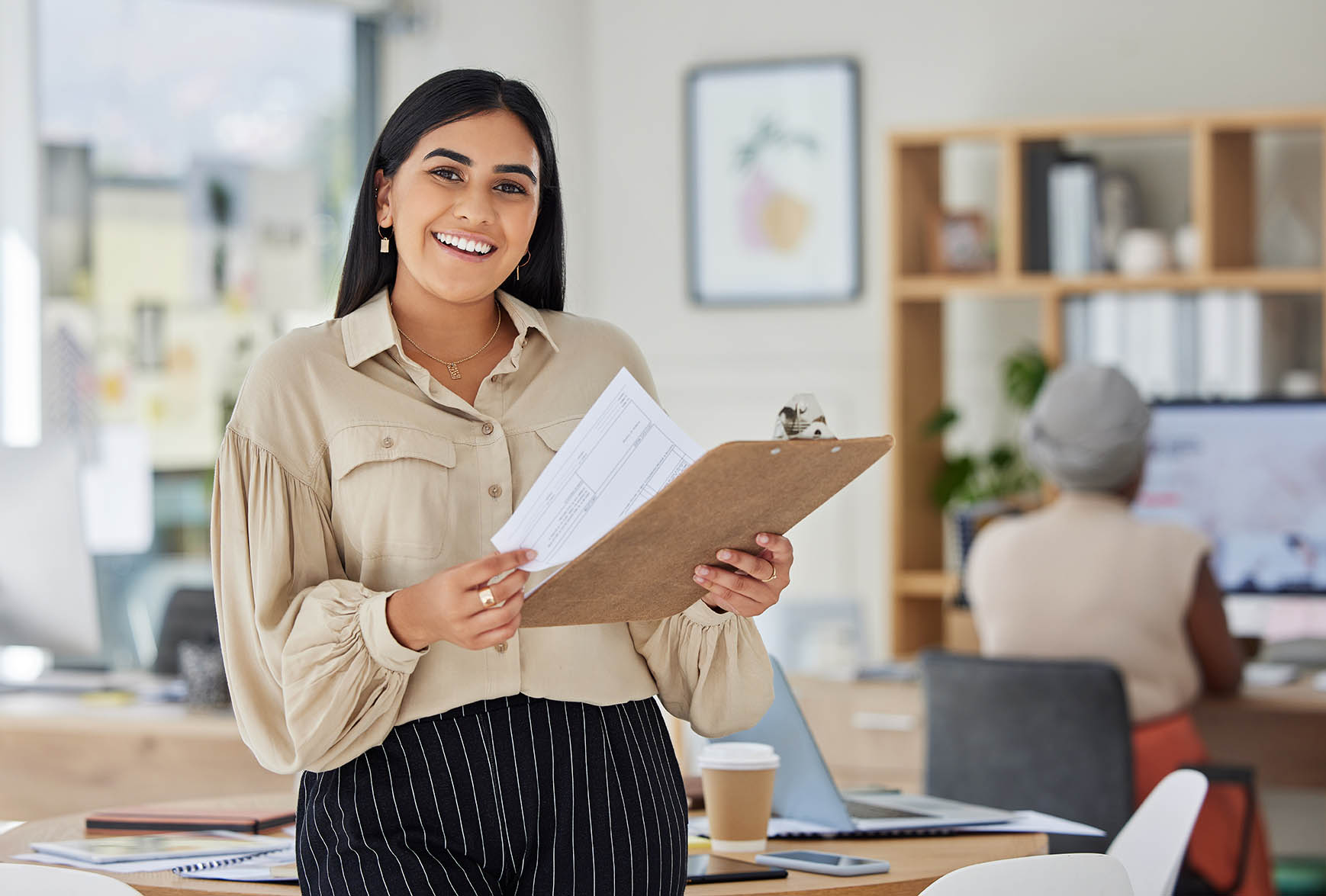 Person standing in an office environment holding a clipboard and several documents. Desks, shelving, office supplies, and another person working at a computer are visible in the background. The image highlights professional support, administration, and workplace collaboration.