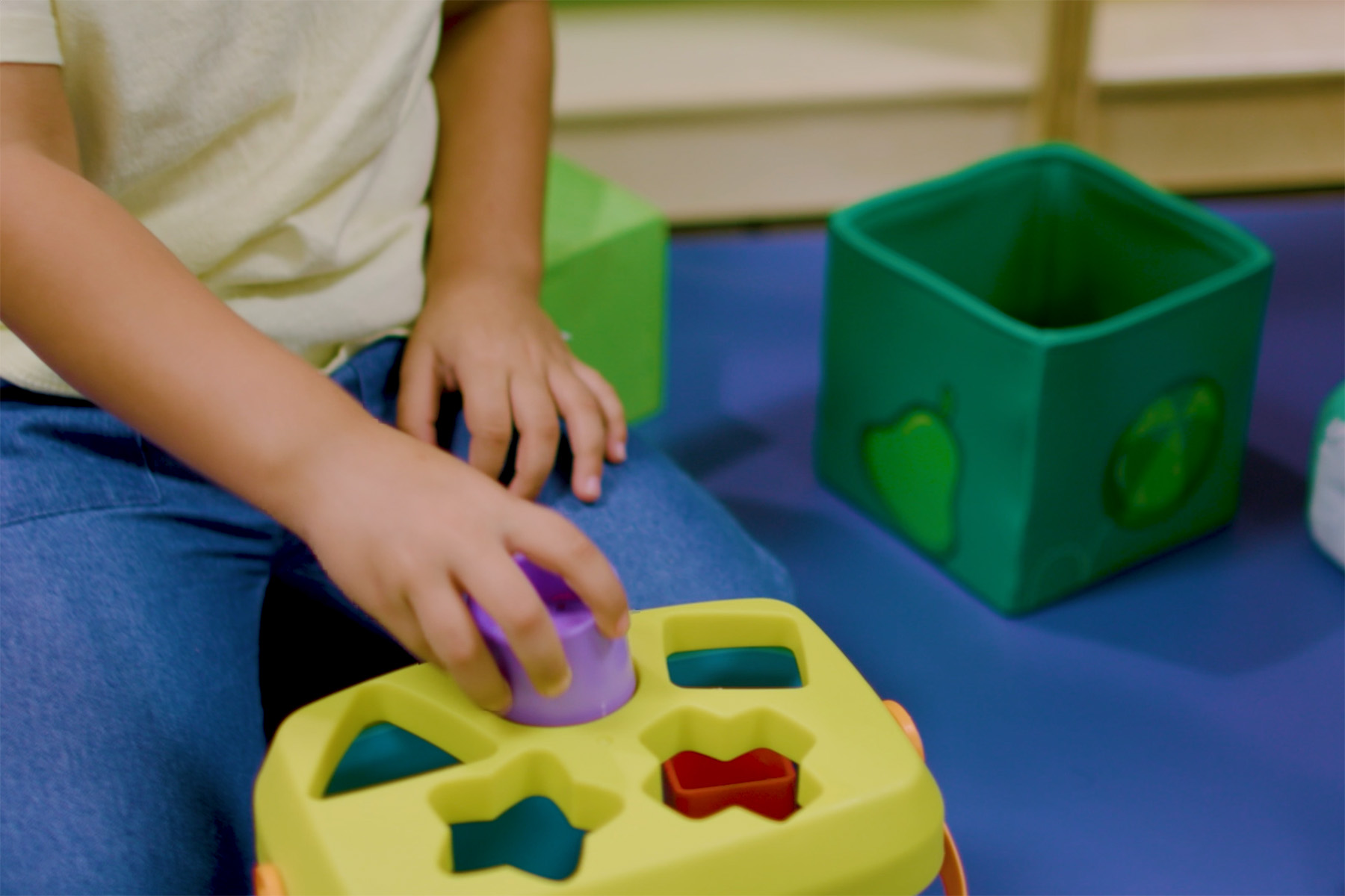 Close-up of a child placing a purple shape into a yellow shape-sorting toy during play. A green storage bin and colorful playroom furnishings are visible in the background, highlighting a hands-on learning activity.