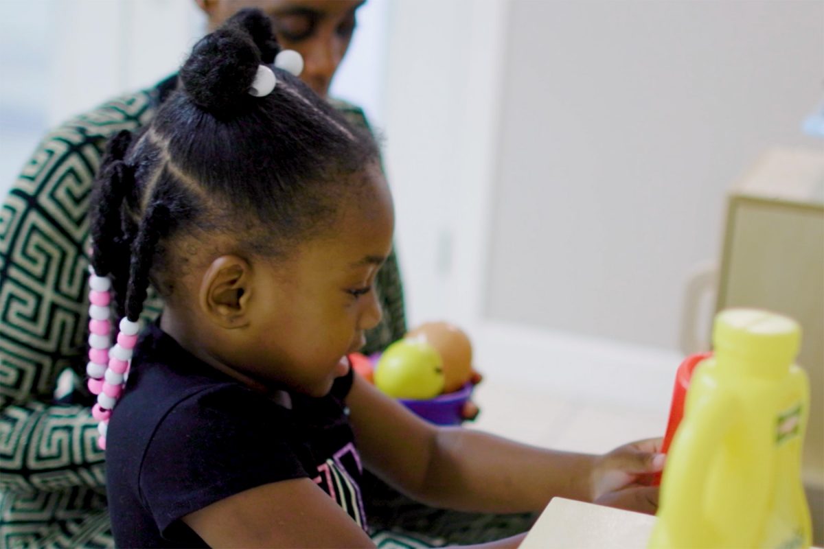 Child engaged in a play-based activity at a table, reaching toward colorful toys and materials. Another person and additional play items are visible in the background, creating a learning and exploration setting.