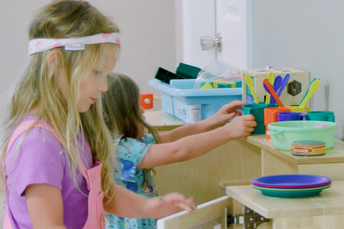 Two children participate in a classroom activity area. One child stands at a shelf selecting materials while another child reaches toward containers of learning supplies. Colorful plates, craft items, and storage bins are arranged on the shelves, highlighting a hands-on early learning environment.