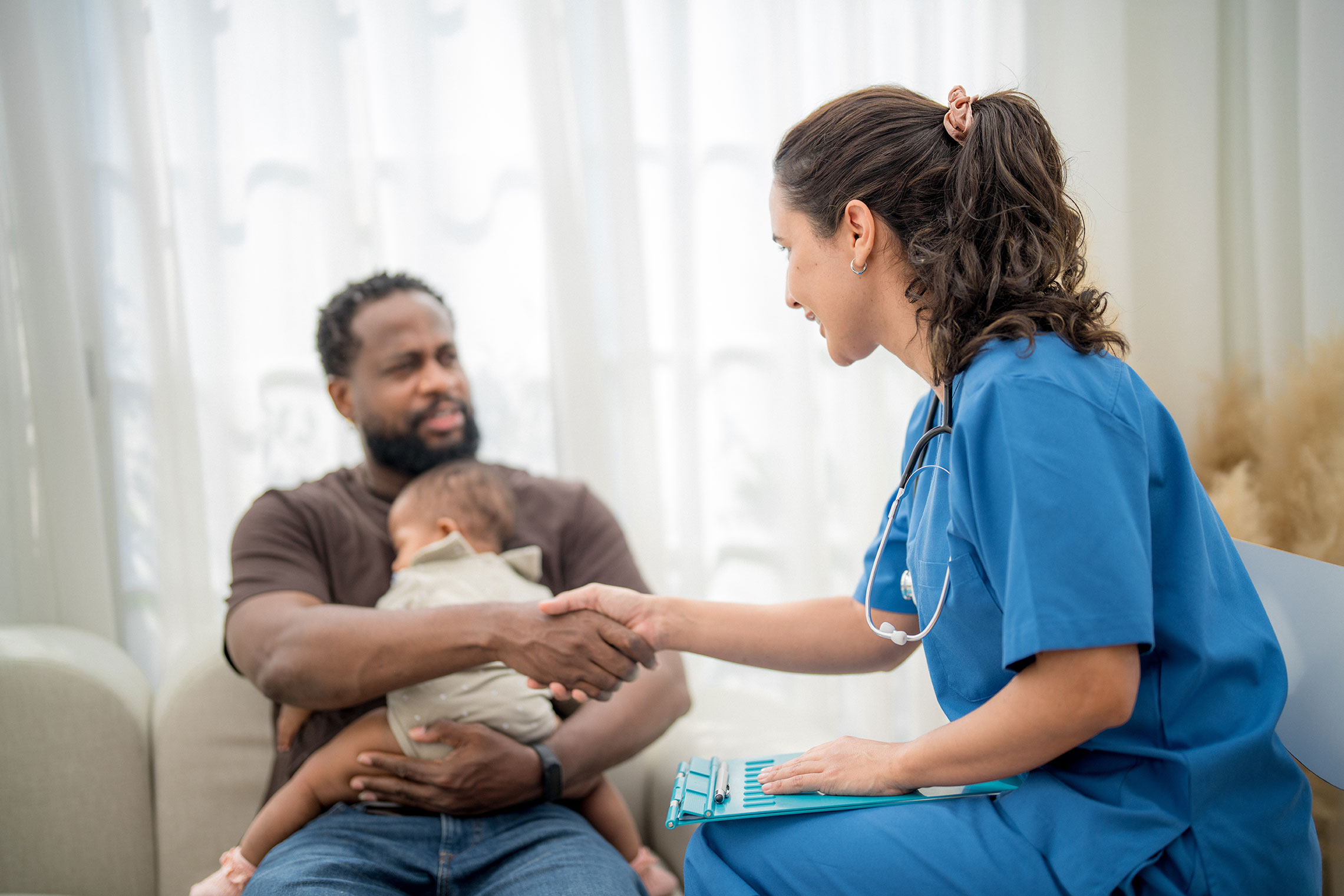 Healthcare provider in blue scrubs shakes hands with a parent holding an infant during a home visit. The healthcare provider holds a clipboard while seated across from the family in a living room setting, highlighting family support, health services, and early childhood care.