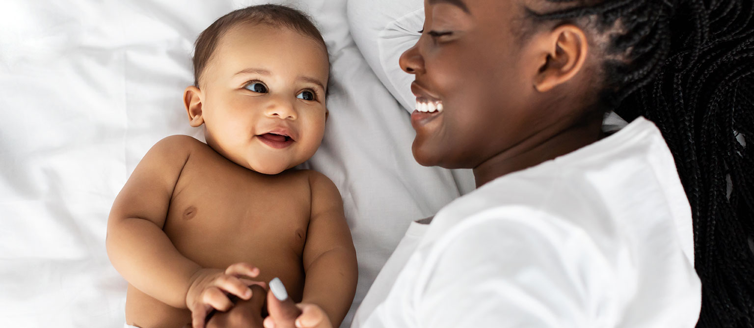 Infant lying on a bed beside a caregiver, with both facing one another during a close interaction. The infant is reaching forward while resting on white bedding, and the caregiver is positioned alongside the infant in a bright indoor setting.