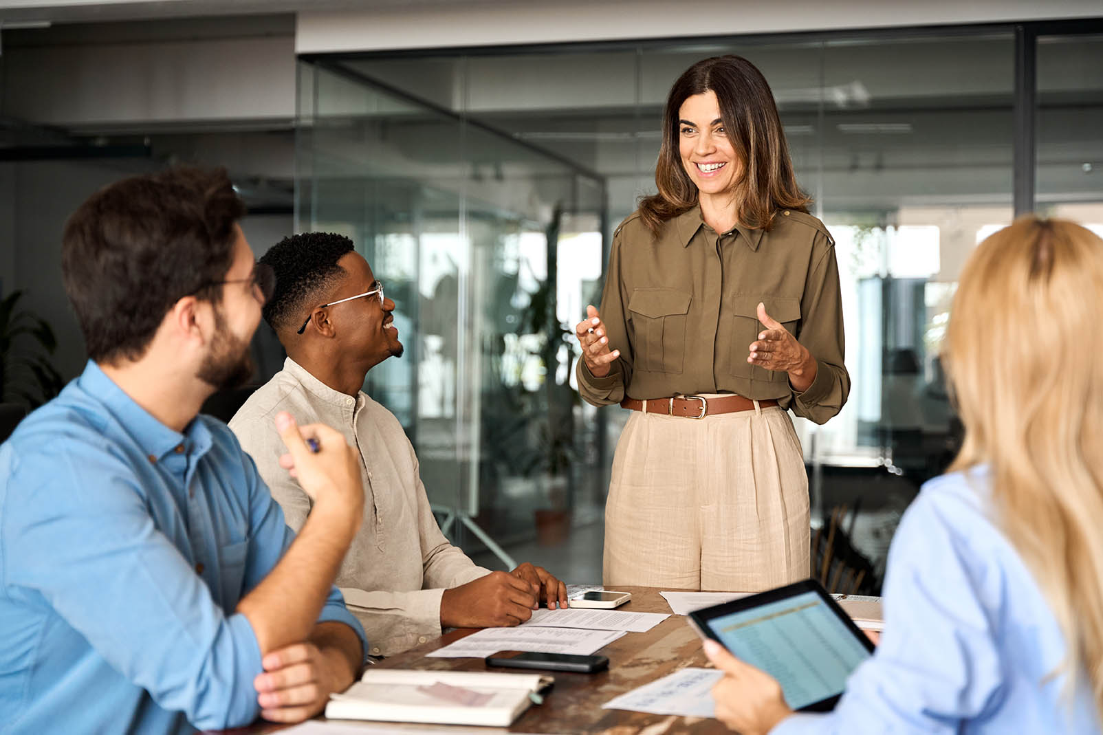 Person stands and presents to a small group during a meeting in a modern office setting. Several participants are seated around a conference table with notebooks, documents, and a tablet computer visible in the foreground. Glass walls and office furnishings appear in the background, highlighting collaboration, leadership, and professional development.