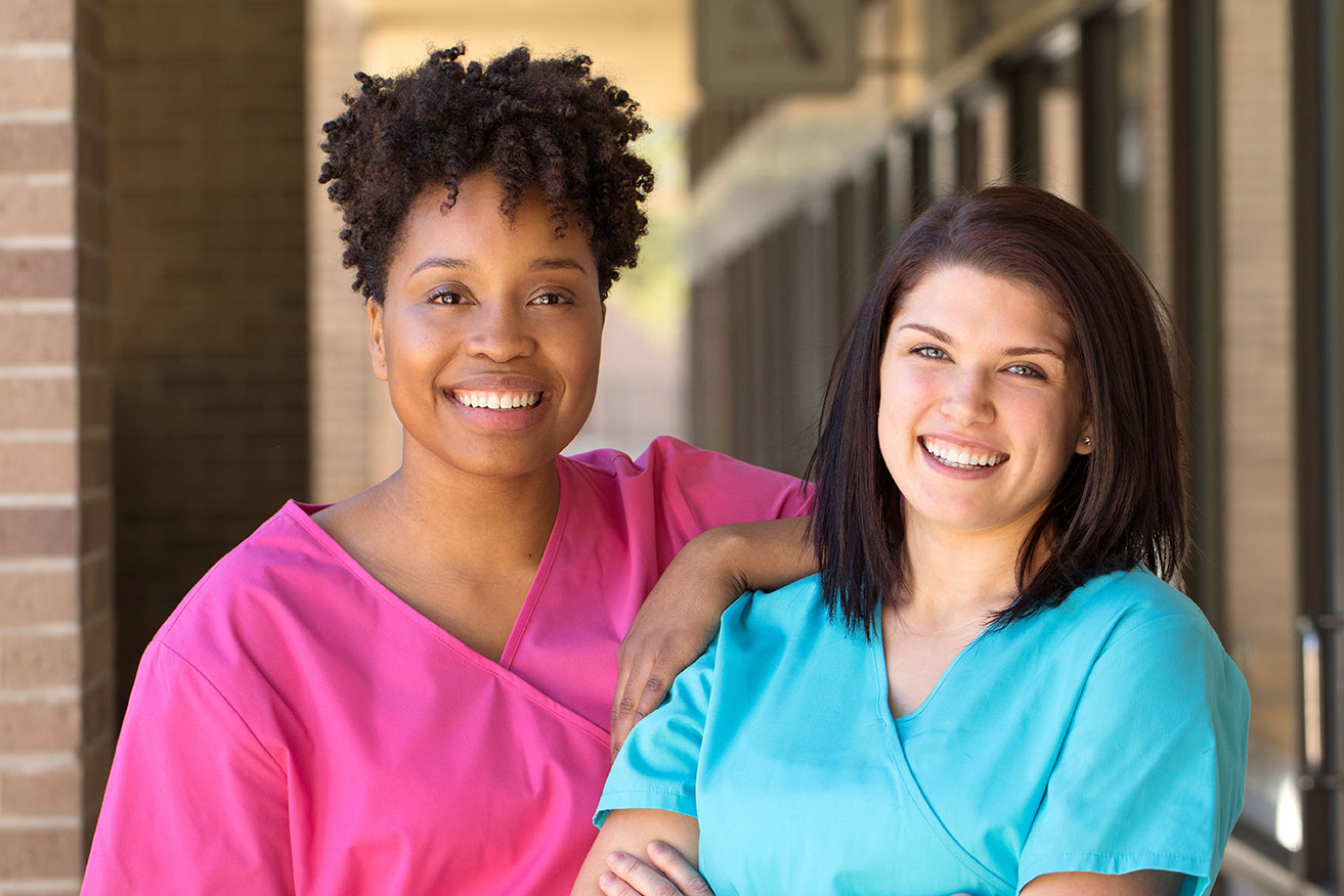 Two healthcare professionals stand side by side outdoors in front of a building. One person wears pink medical scrubs and the other wears blue medical scrubs. The image highlights a professional healthcare setting and supportive service providers.
