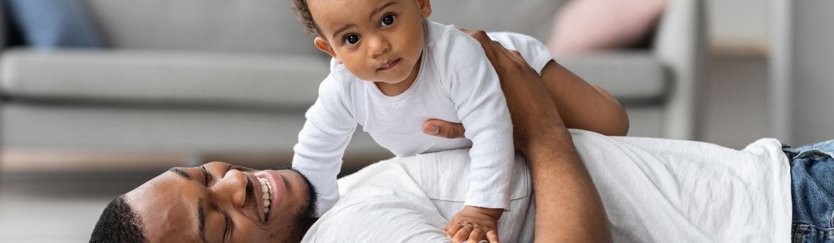 Caregiver lying on the floor while supporting an infant during a play activity. The infant, dressed in a white long-sleeve outfit, is positioned above the caregiver and looking toward the camera. The scene takes place in a home setting with a sofa visible in the background, highlighting positive parent-child interaction and early development.