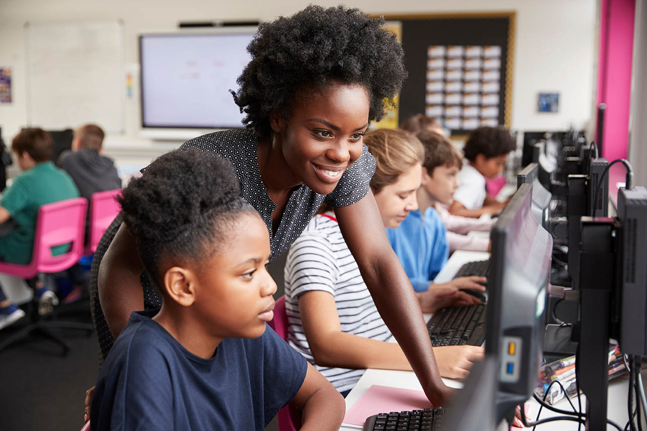 Adult stands beside a child seated at a desktop computer in a classroom computer lab. Several children work at computers arranged in rows, while the adult provides guidance and support. Classroom furnishings, computer monitors, and educational materials are visible in the background, highlighting technology-based learning and instructional support.