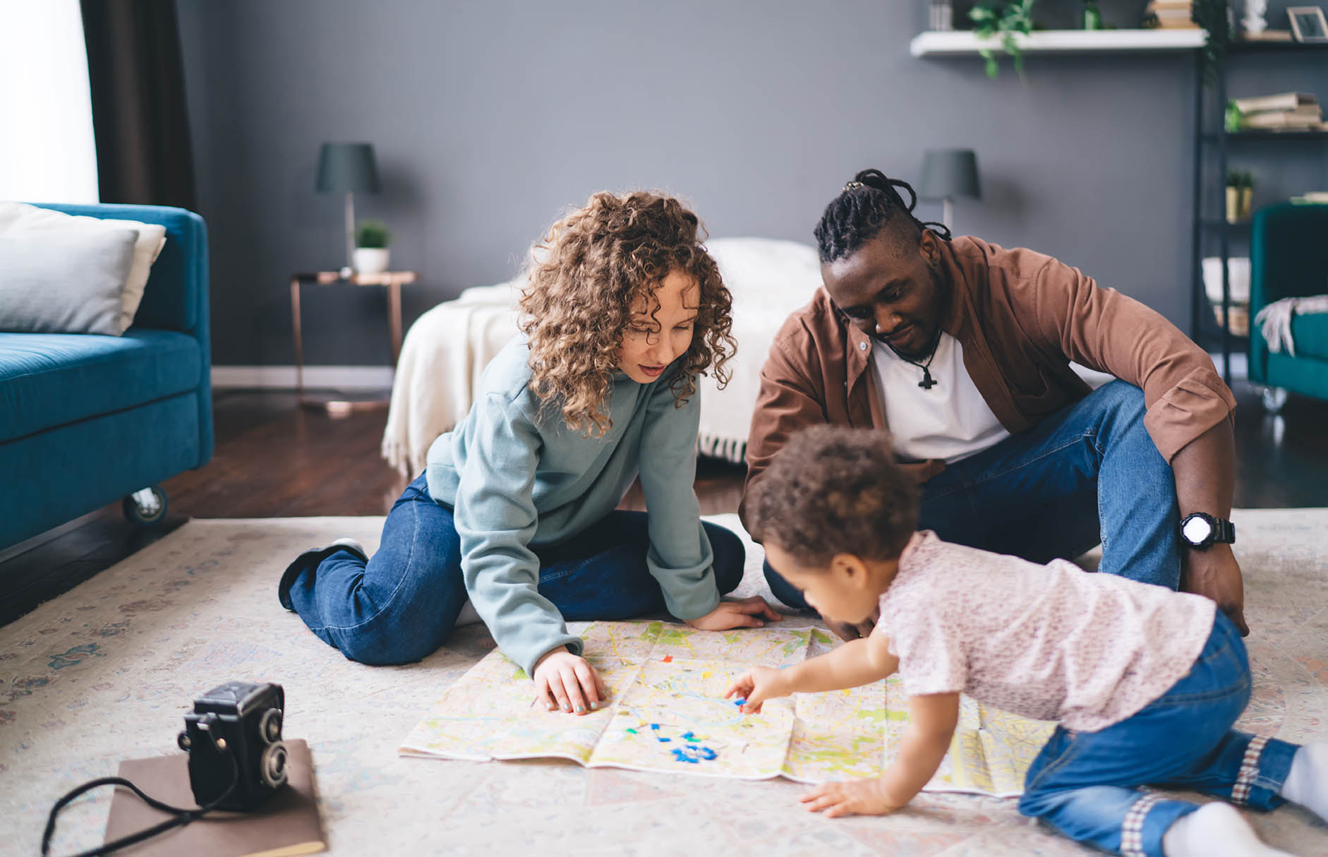 Two adults and a young child gather on the floor of a home while looking at a large map or activity sheet. The child crawls across the map as the adults kneel nearby and participate in the activity. A sofa, bed, and camera are visible in the background, highlighting family engagement, learning, and interactive play in a home environment.