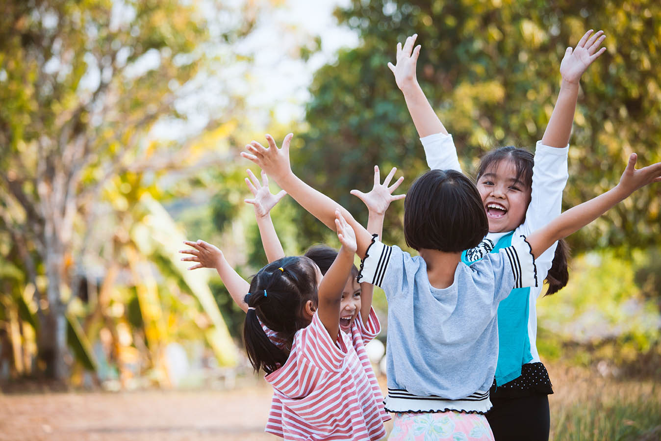 Group of children playing together outdoors with their arms raised while standing in a circle. Trees and greenery surround the play area, and sunlight filters through the background. The image highlights active play, social interaction, and outdoor recreation in a natural setting.