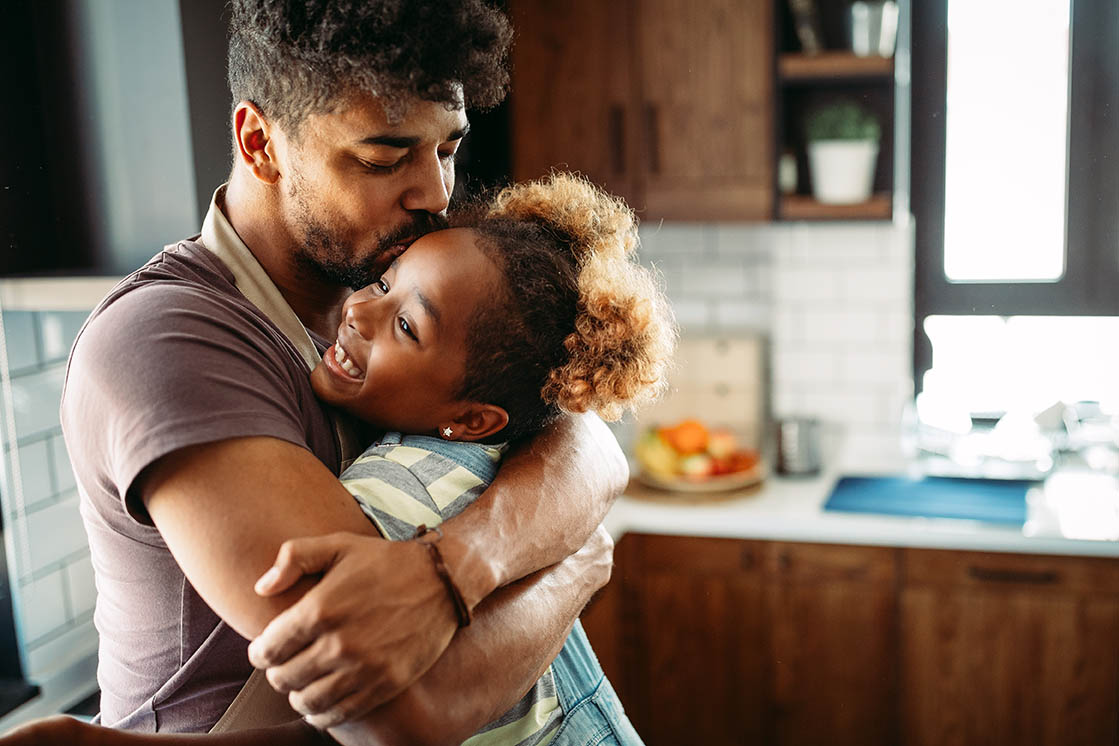 Adult embraces a young child in a kitchen setting. The child rests in the adult’s arms while household cabinets, countertops, and kitchen items are visible in the background. The image highlights a close caregiver-child interaction in a home environment.