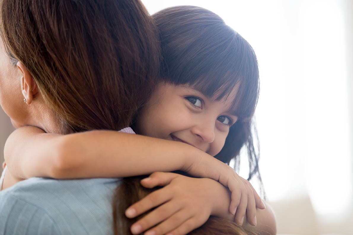 Young child embraces an adult, with the child’s arms wrapped around the adult’s shoulders. The child looks toward the camera while resting against the adult. Soft natural light and a neutral indoor background create a warm home setting, highlighting a close caregiver-child interaction.