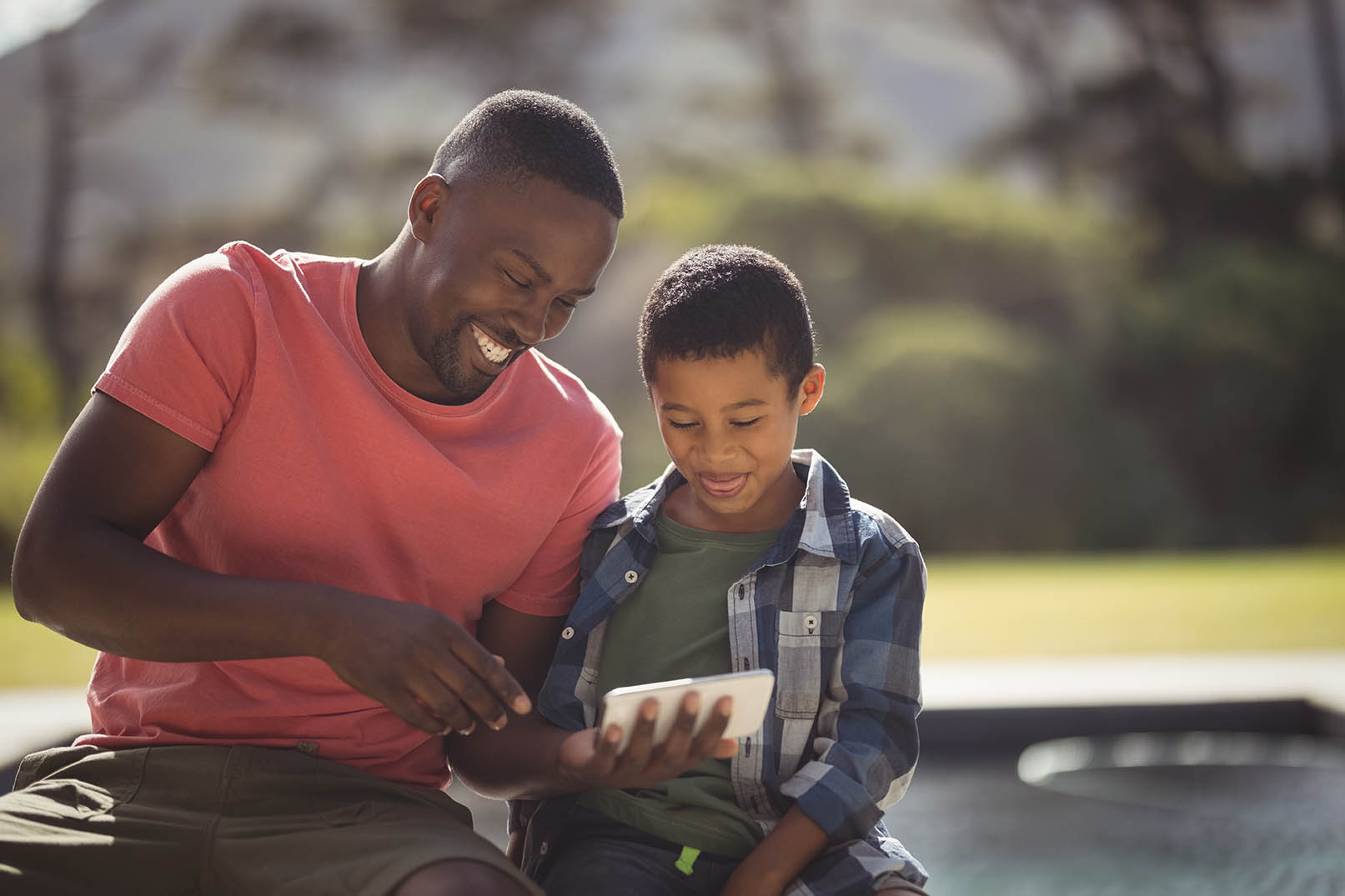 Adult and child sit together outdoors looking at a tablet device. The adult points toward the screen while the child holds the tablet. Trees and greenery are softly blurred in the background, highlighting shared learning, technology use, and caregiver-child engagement in an outdoor setting.