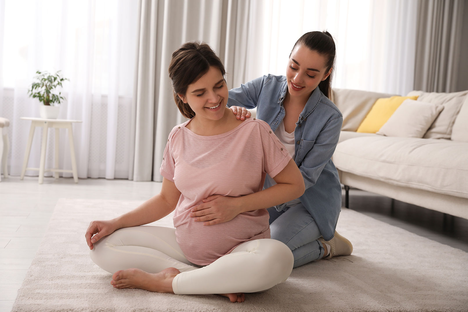 Pregnant individual seated on a rug in a home setting while receiving physical support from a healthcare provider or support professional. The support professional places one hand on the individual's shoulder and another near the lower back as they practice a seated stretching or comfort technique. A sofa and soft furnishings are visible in the background, highlighting prenatal care and maternal wellness support.