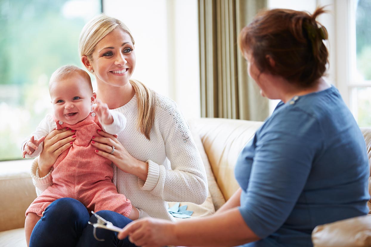 "Person holding a baby while talking with another adult during a home visit."