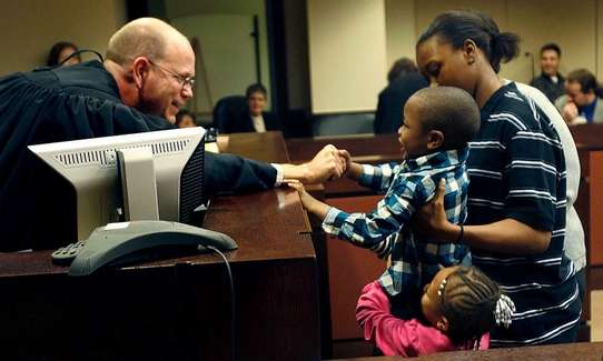 Judge reaches across a courtroom bench to shake hands with a child standing beside an adult, while another child stands nearby. Courtroom furnishings, including a computer monitor and seating area, are visible in the background, highlighting a family-centered judicial setting and positive engagement between the court and families.
