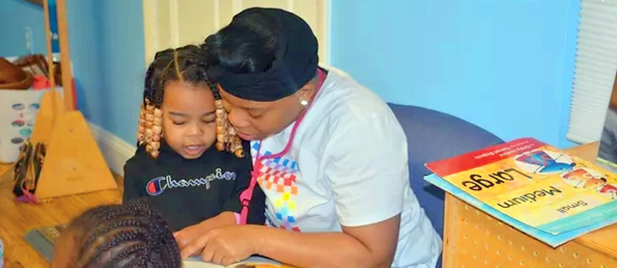 Adult and two children participate in a shared reading activity in a classroom setting. The adult points to a book while a child sits nearby, and additional children's books are displayed on a shelf beside them, emphasizing literacy and early learning.