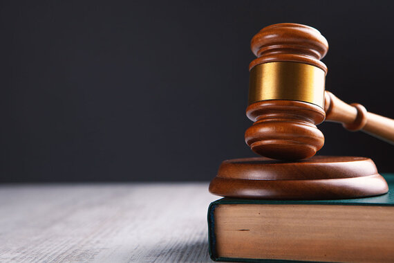 Close-up of a wooden judge’s gavel resting on a sound block atop a thick law book. The image is set against a dark background with ample open space, symbolizing the legal and judicial system.