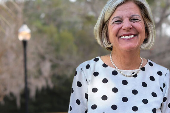 Portrait of Dr. Mimi Graham outdoors, wearing a light-colored blouse with black polka dots and a pearl necklace. Trees and a lamp post appear softly blurred in the background, creating a professional portrait setting.